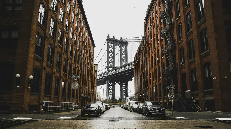 A cobblestone street lined with red brick buildings leads toward the Manhattan Bridge on an overcast day, with parked cars along both sides