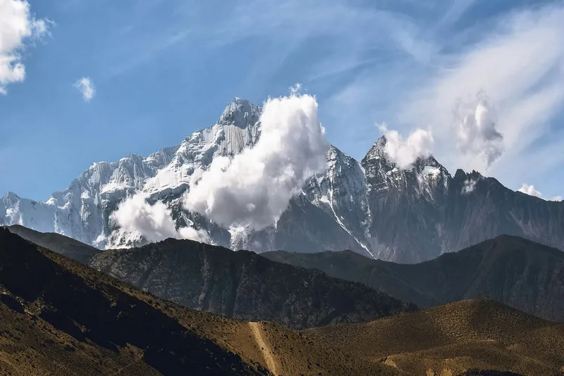 Snow-covered mountain peaks rise above dark hills under a bright blue sky with drifting clouds