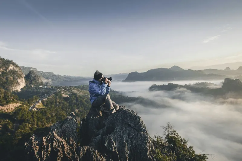 A photographer sits on a rocky cliff overlooking a misty mountain valley while taking a picture at sunrise
