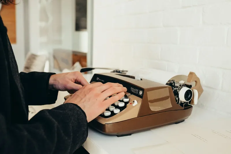 A person types on a vintage typewriter placed on a white desk beside a brick wall