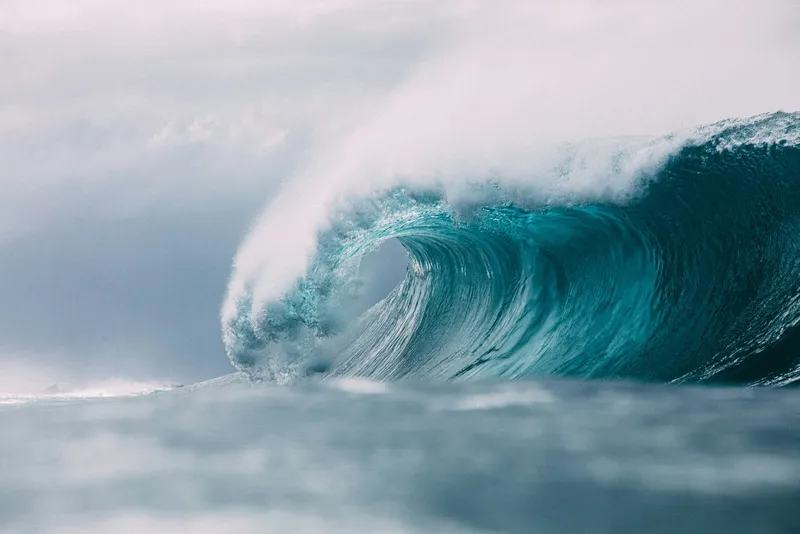 A massive turquoise ocean wave curls into a hollow barrel under a cloudy sky, with sea spray misting across the water